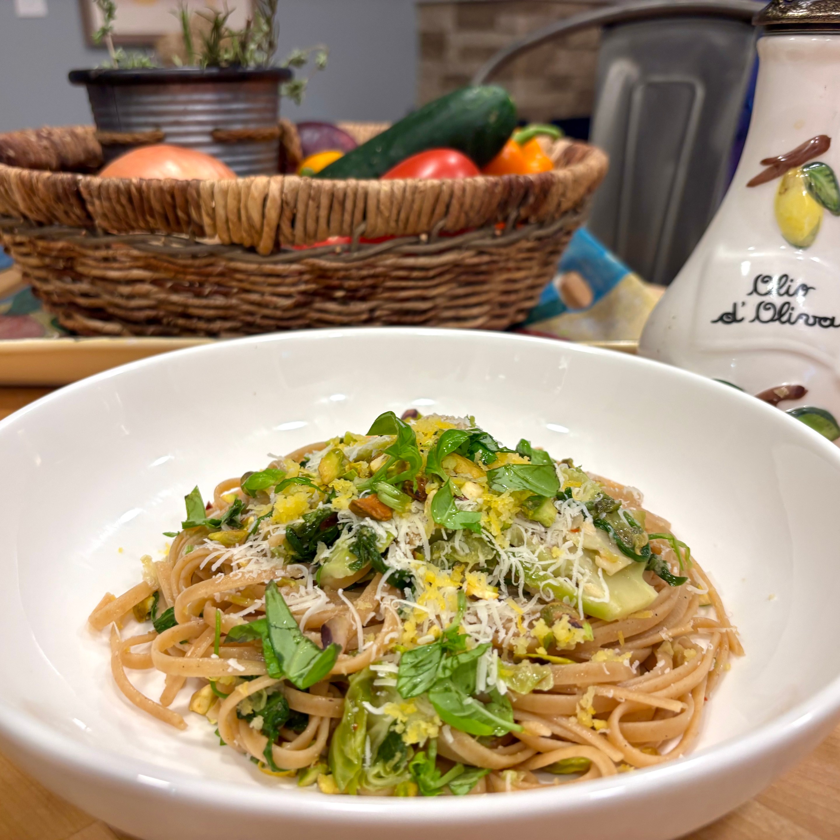 A bowl of whole grain pasta with vegetables, pistachios, and cheese on a kitchen table with a basket of produce in the background.