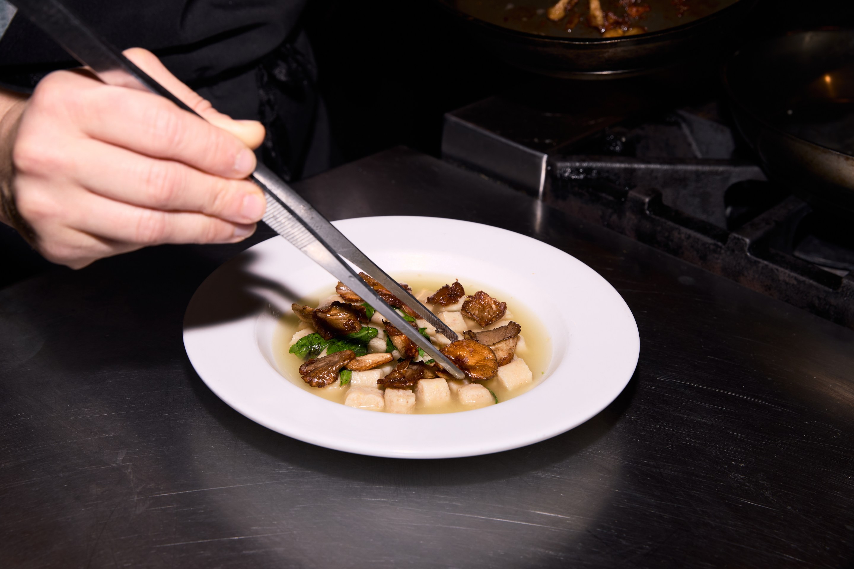 Passatelli pasta in broth with oyster mushrooms and shaved parmesan, served in a white bowl on a black table.