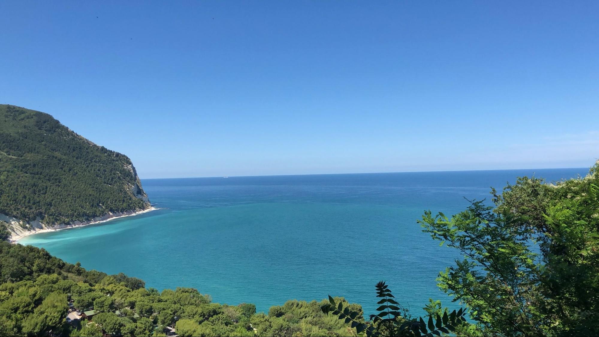 Picturesque view of Sirolo, Italy, with blue bay in distance, surrounded by hills and lush vegetation.