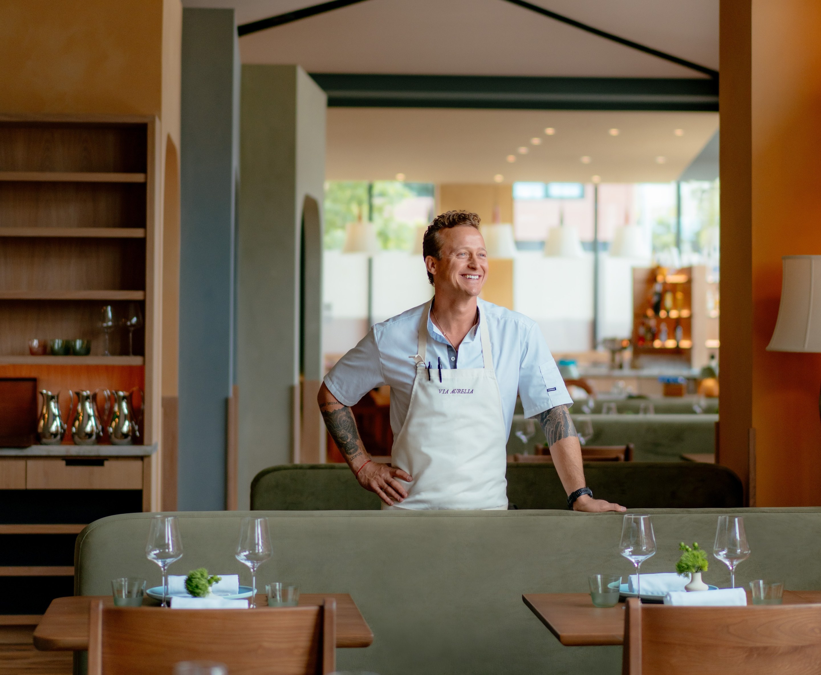 Chef David Nayfeld of Via Aurelia, standing in apron inside the dining room, surrounded by set tables and olive couches.