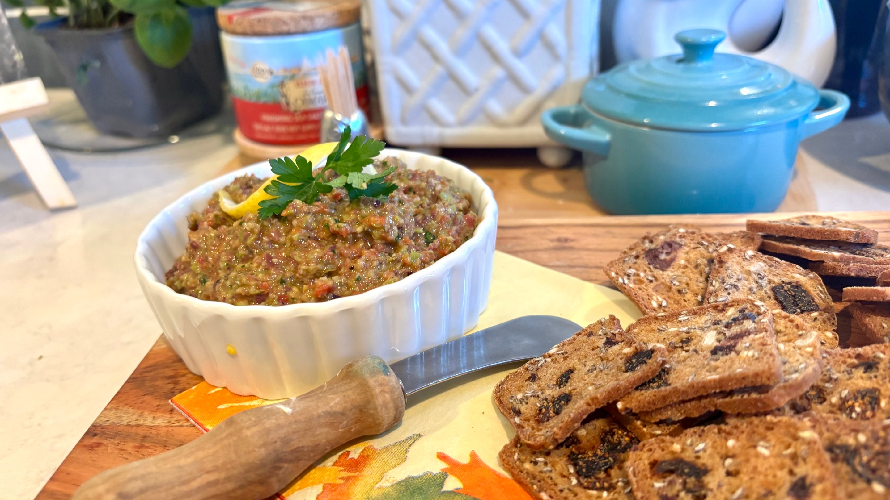 Bowl of Sicilian tapenade garnished with lemon and parsley beside rustic fig crackers on a wooden board.