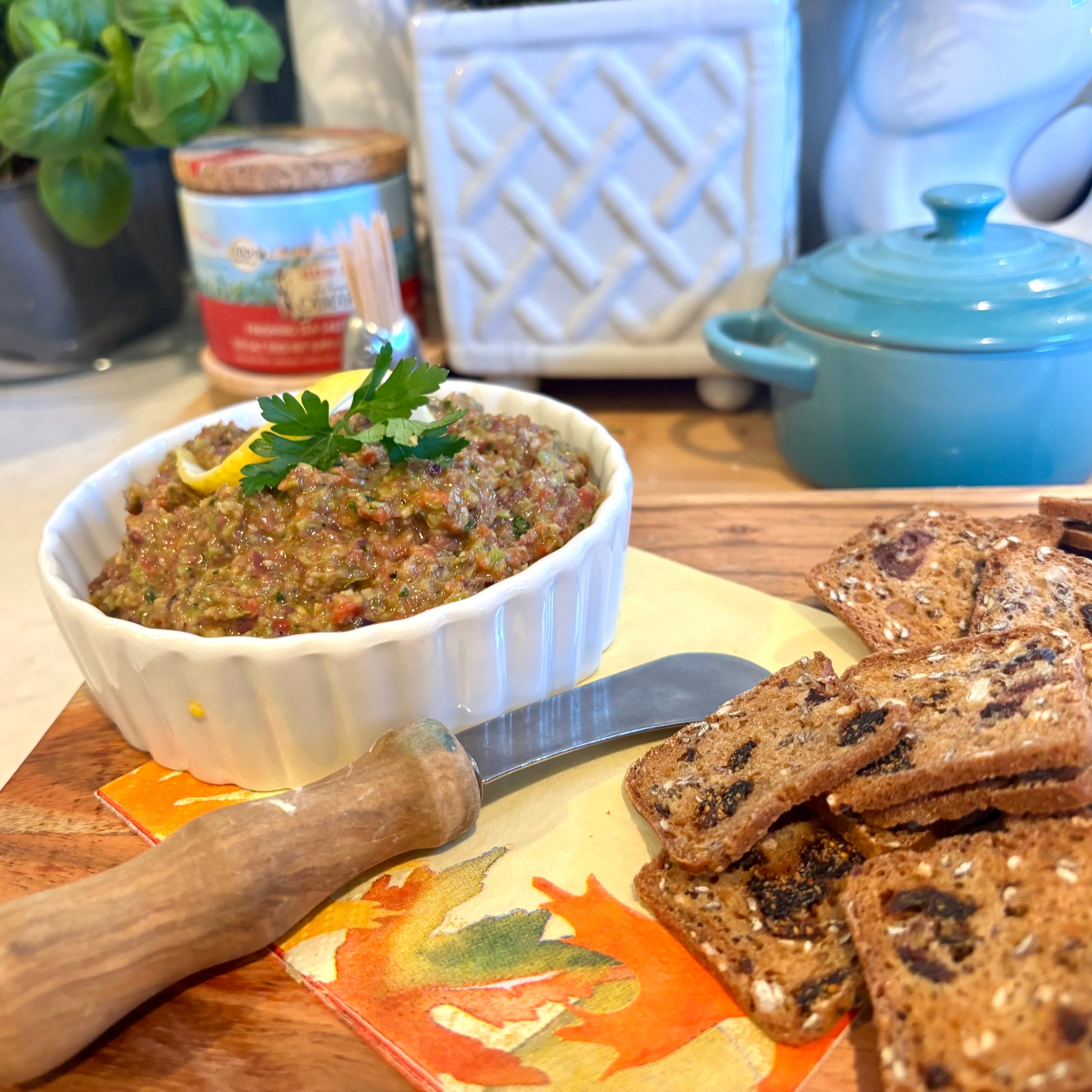 Bowl of Sicilian tapenade garnished with lemon and parsley beside rustic fig crackers on a wooden board.