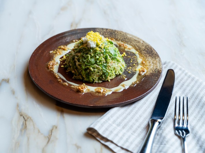 Brussels sprout salad with Meyer lemon dressing and Marcona almond pesto at Bottega Napa Valley., served on a round brown plate on a marble table.