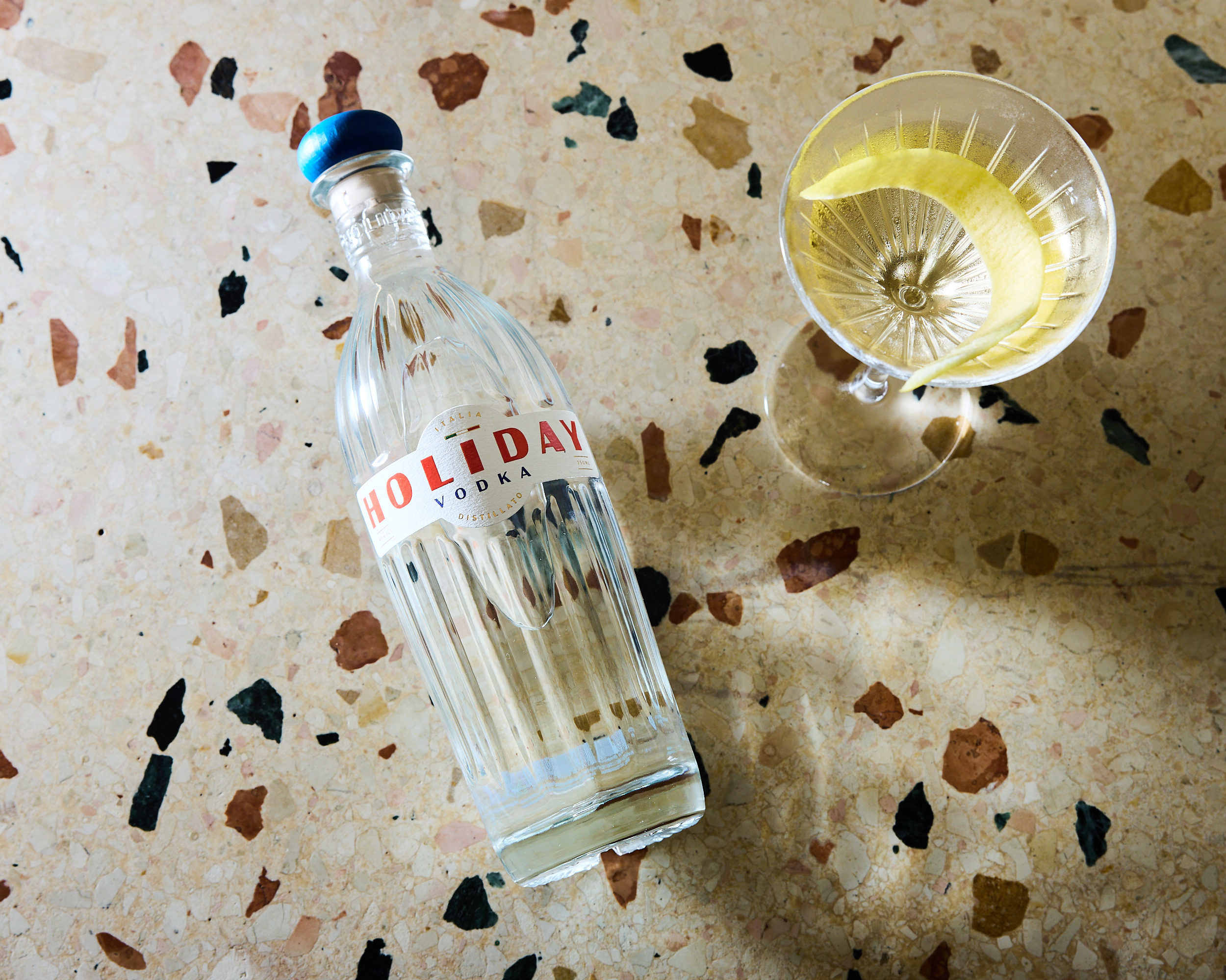 Holiday Vodka bottle on terrazzo surface beside cocktail with lemon twist, symbolizing Italian elegance and modern design.