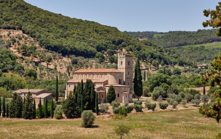 Romanesque abbey surrounded by cypress trees and Tuscan hills.