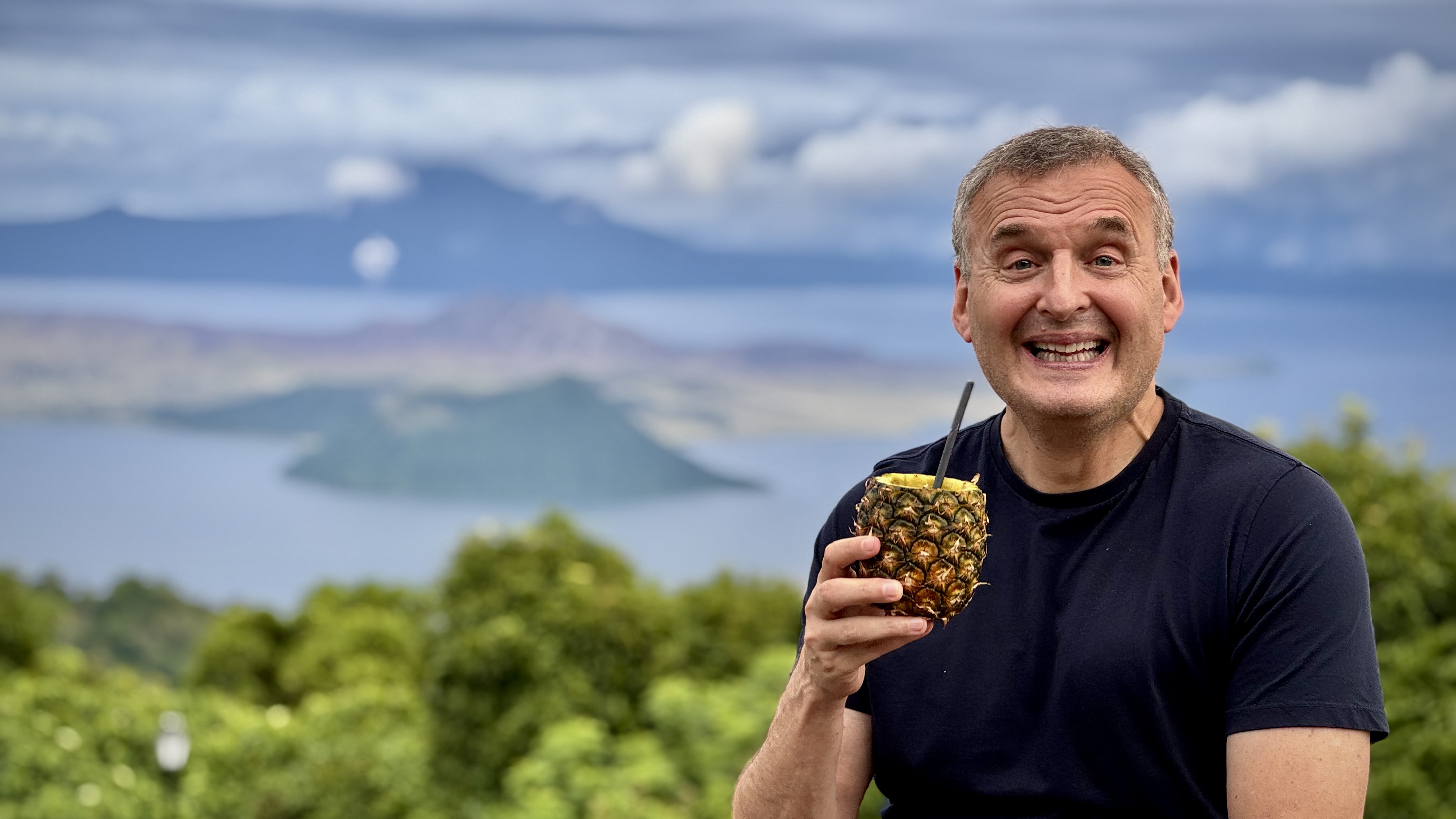 Phil Rosenthal smiles while enjoying a pineapple drink with a tropical view behind him.