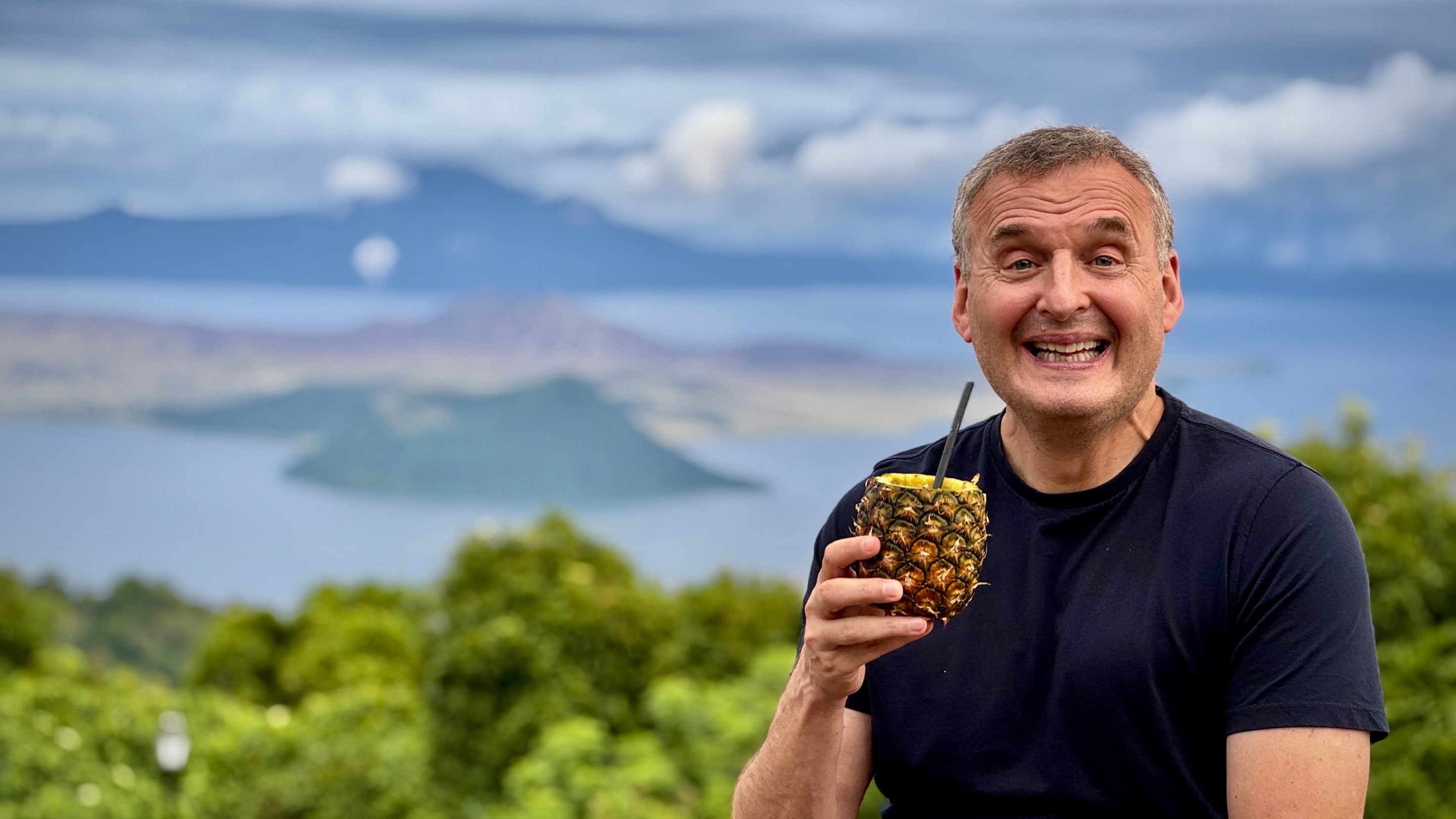 Phil Rosenthal smiles while enjoying a pineapple drink with a tropical view behind him.