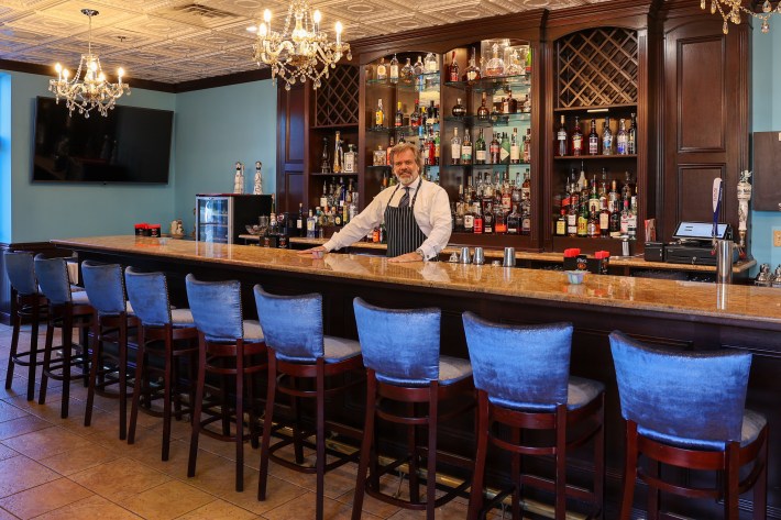Marble bar with chandeliers, blue velvet barstools, and wood cabinetry inside Vette Ristorante in Livingston NJ.