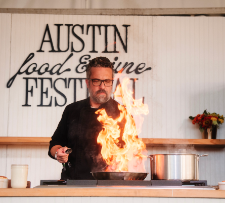 Fabio Viviani standing at the demo counter with a flaming pan during the Austin Food and Wine Festival.