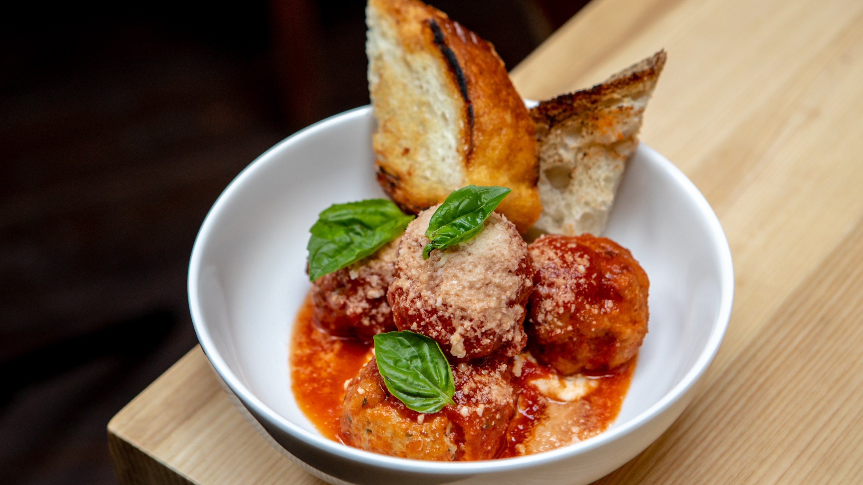 ‘Ino’s Red Wattle Pork Meatballs with tomato sauce and ricotta served in a white bowl on a wooden cutting board.