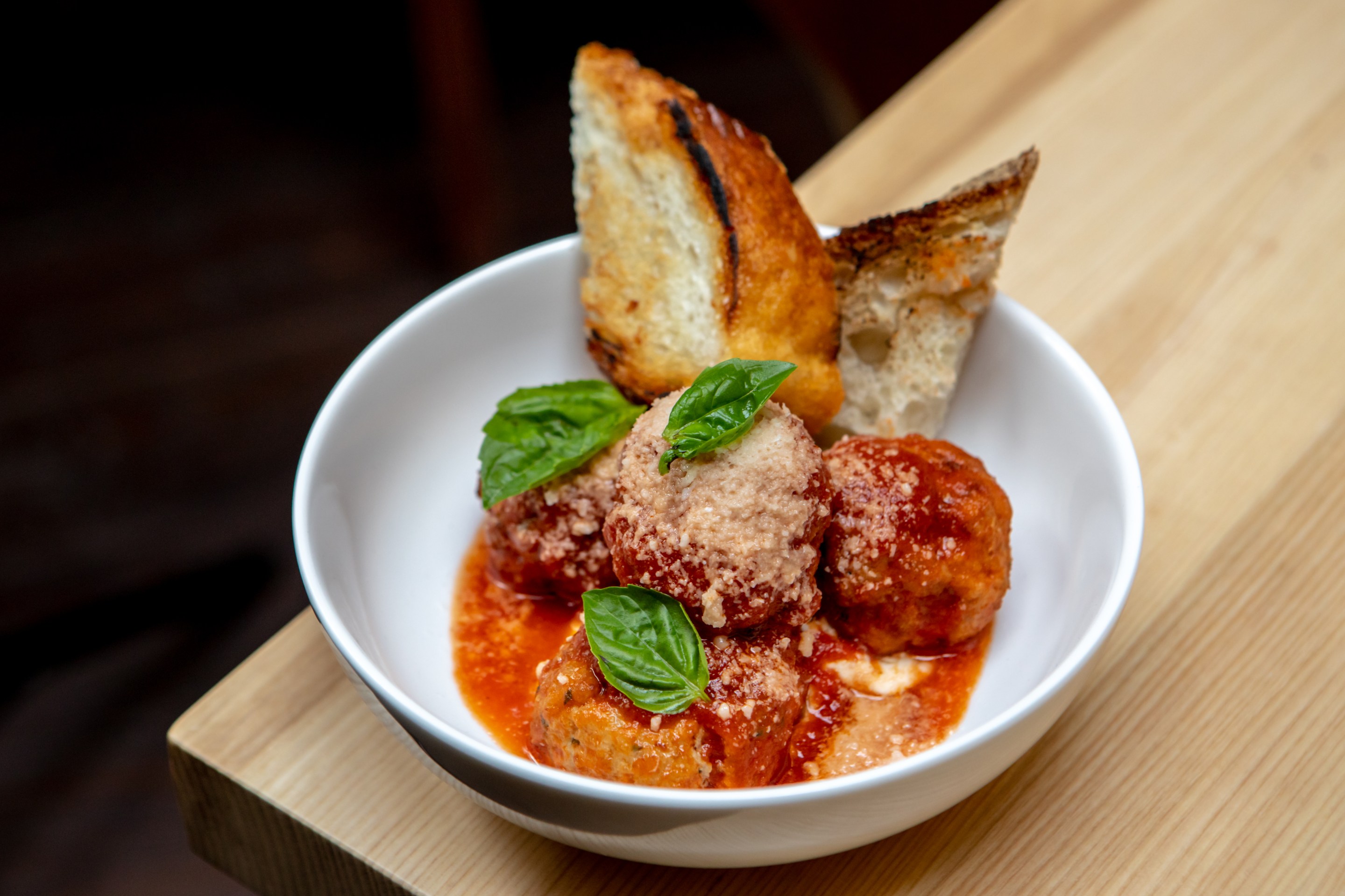 ‘Ino’s Red Wattle Pork Meatballs with tomato sauce and ricotta served in a white bowl on a wooden cutting board.