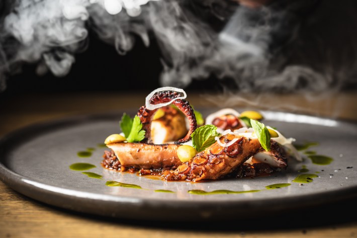 Charred octopus plated with nduja sugo, smoked potato, lemon, and fresh herbs.