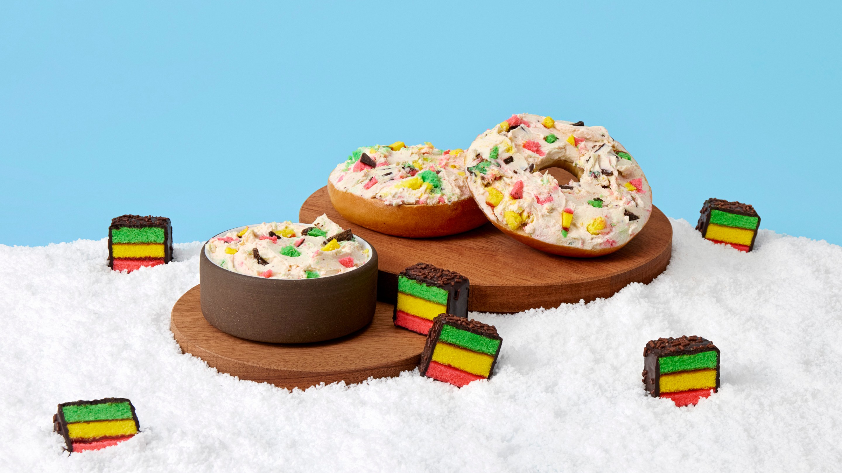 Bagels and cream cheese mixed with rainbow cookie pieces on a snowy white surface with scattered mini rainbow cookies.