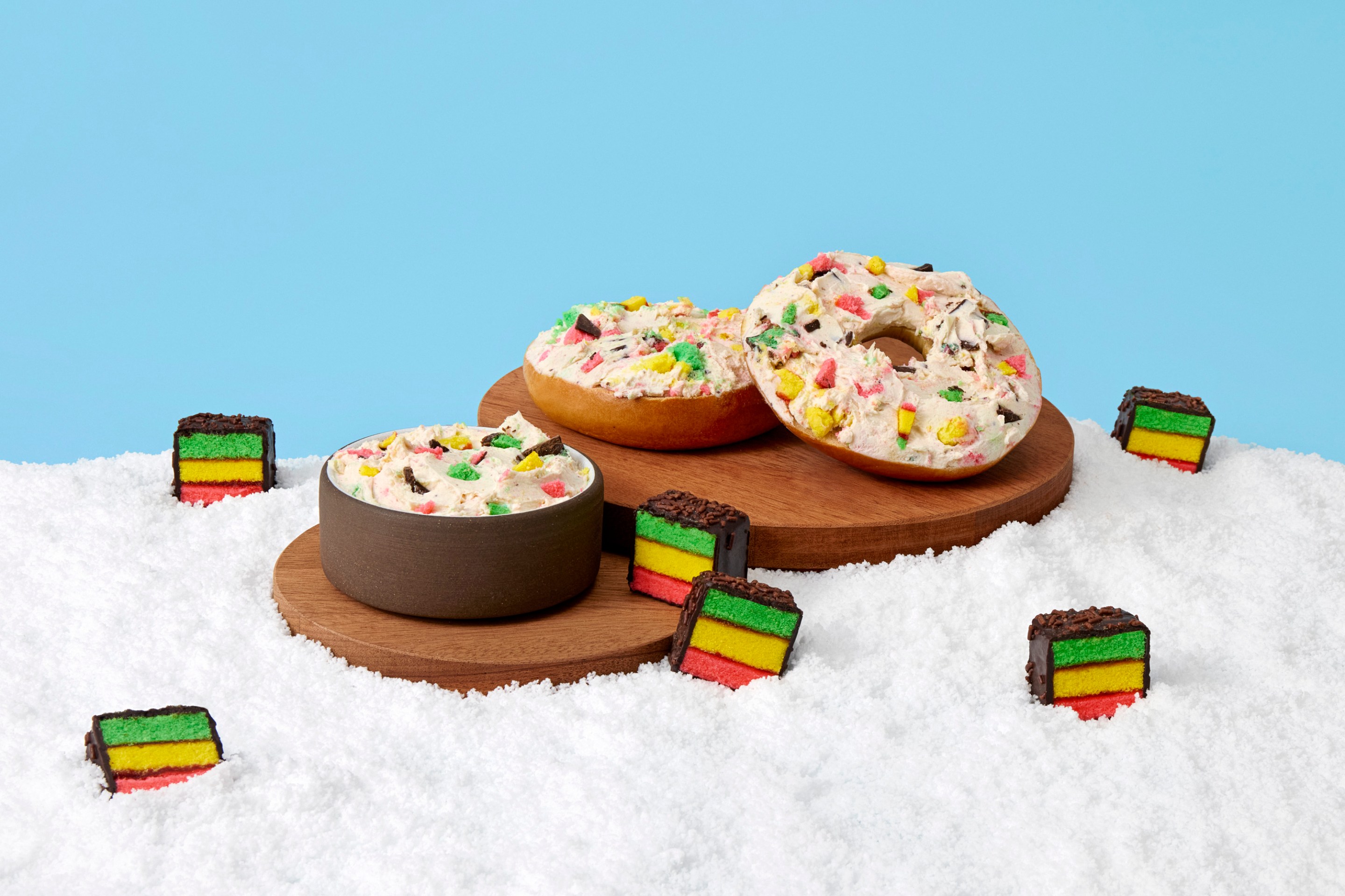 Bagels and cream cheese mixed with rainbow cookie pieces on a snowy white surface with scattered mini rainbow cookies.