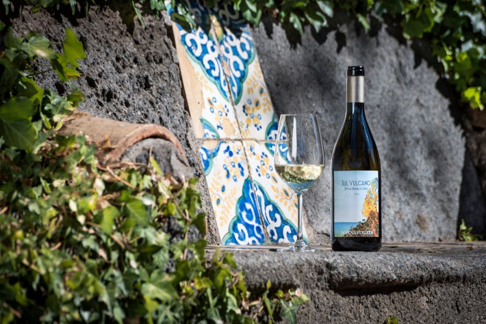 Bottle of Sul Vulcano Etna Bianco with a wine glass placed on volcanic rock near Sicilian ceramic tiles.