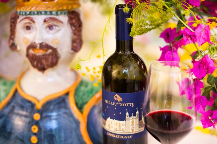 Bottle of Mille e Una Notte with glass of red wine and Sicilian ceramics in the background.