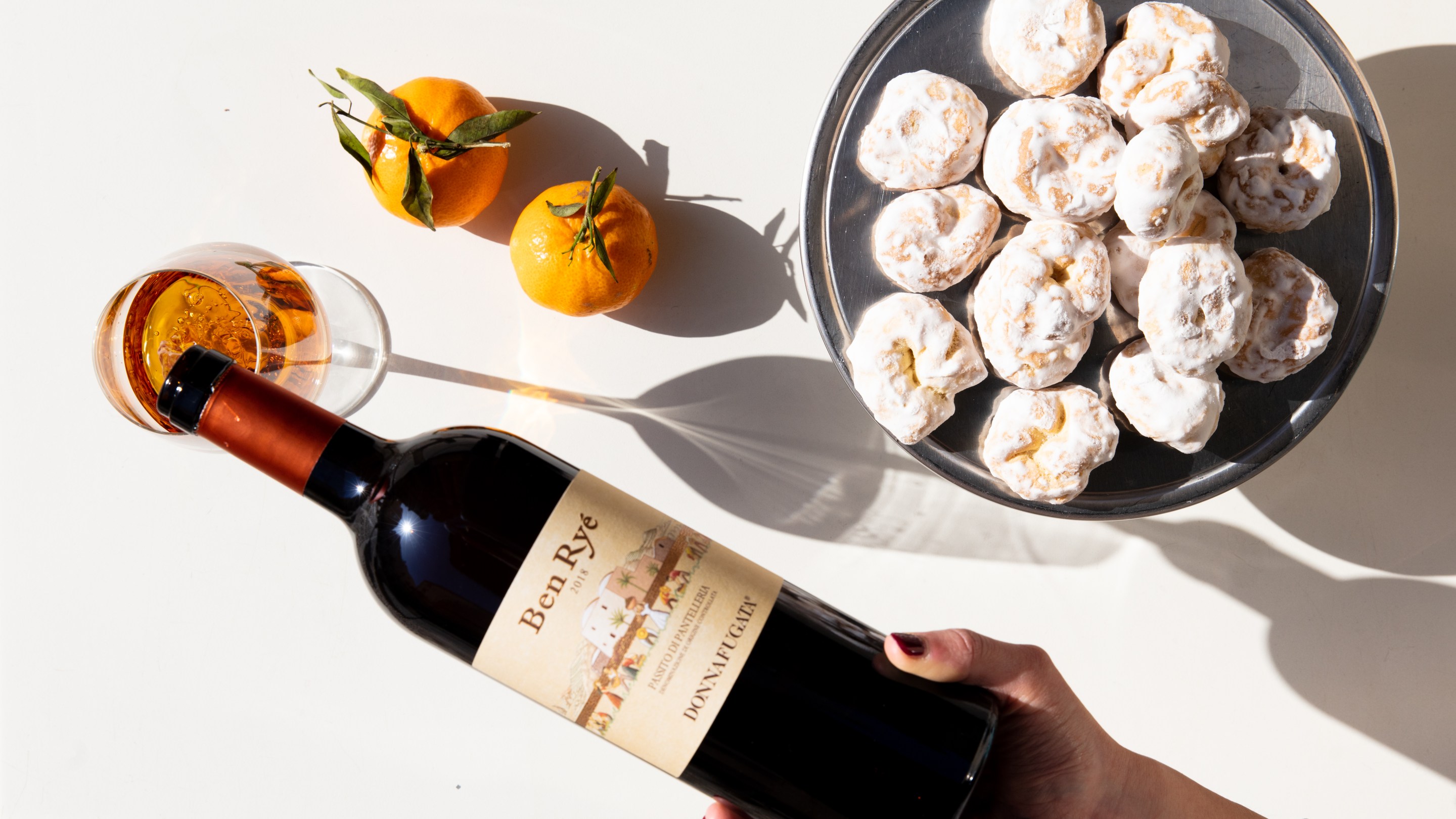 Bottle of Ben Ryé Passito di Pantelleria with citrus fruits, pastries, and a glass of dessert wine.