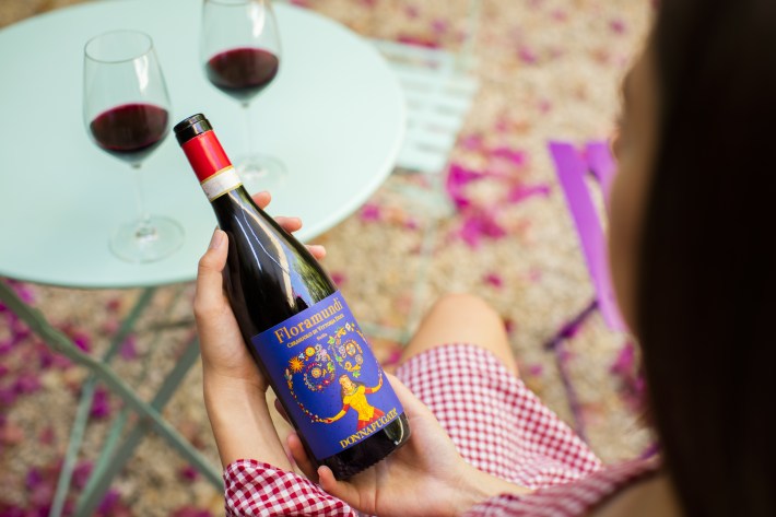 Person holding a bottle of Floramundi wine with two glasses of red wine on a table outdoors.