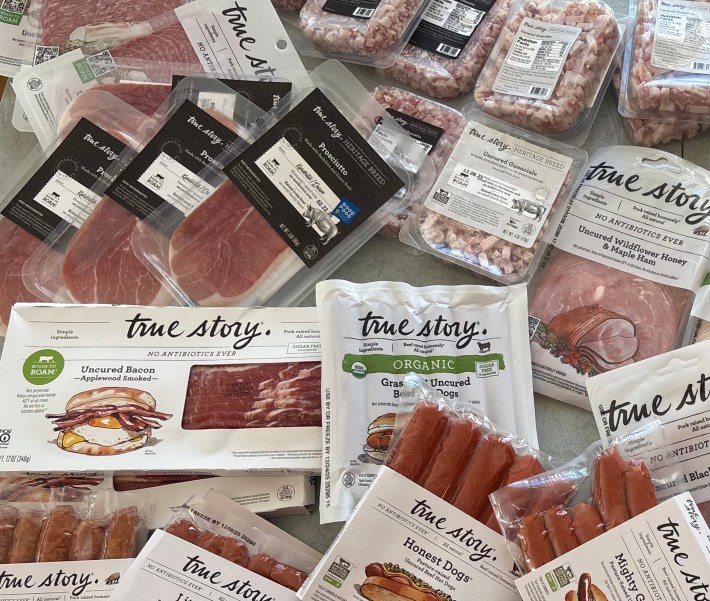 Assorted True Story Foods packages of prosciutto, ham, bacon, sausage, and ground pork laid out on a counter.