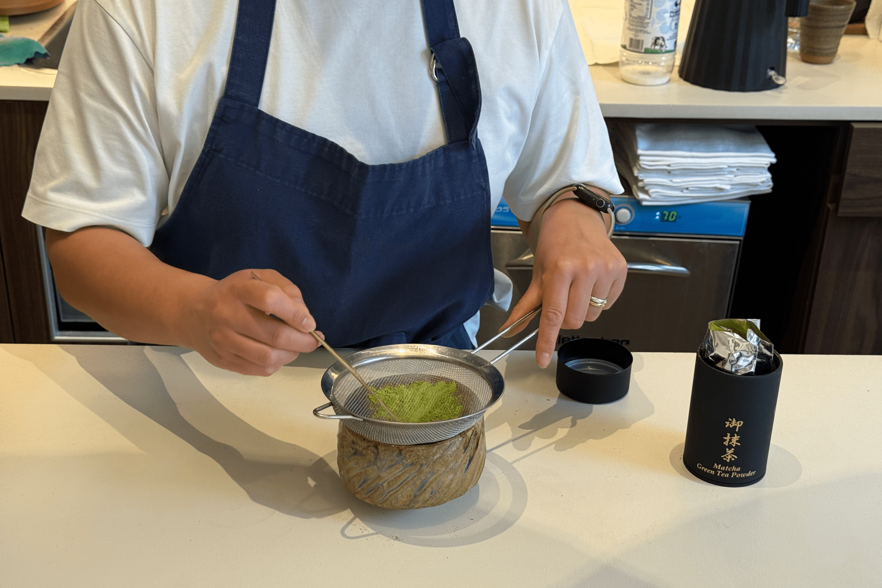 Barista hand-whisking matcha through a sieve into a ceramic bowl at Emoraya Shop in Milan.