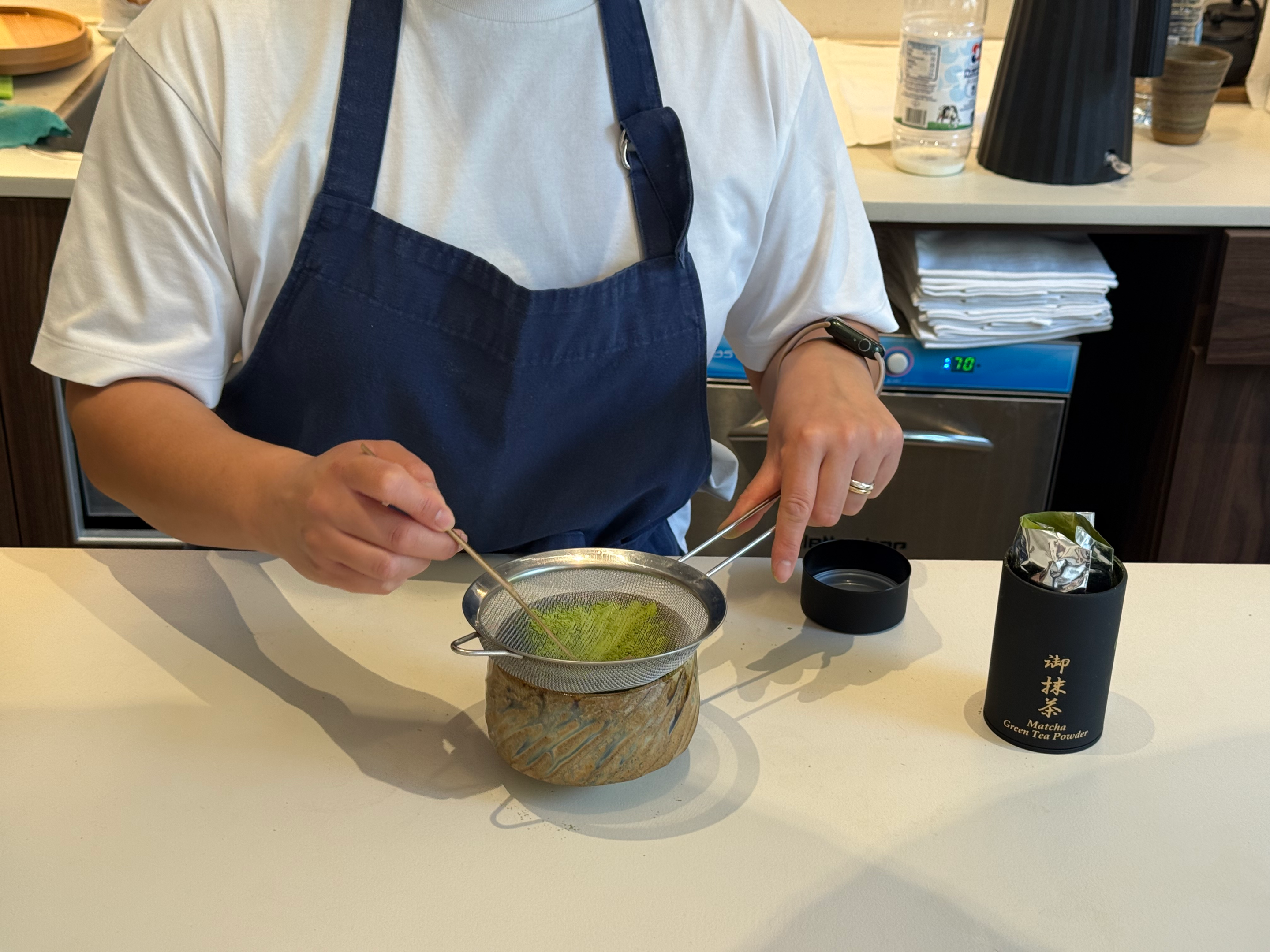 Barista hand-whisking matcha through a sieve into a ceramic bowl at Emoraya Shop in Milan.