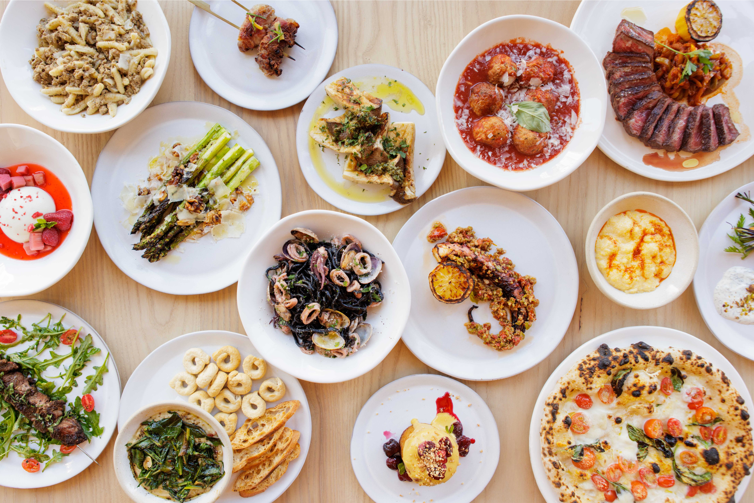 Italian food in white bowls on a light wooden table - from meatballs to pizza.