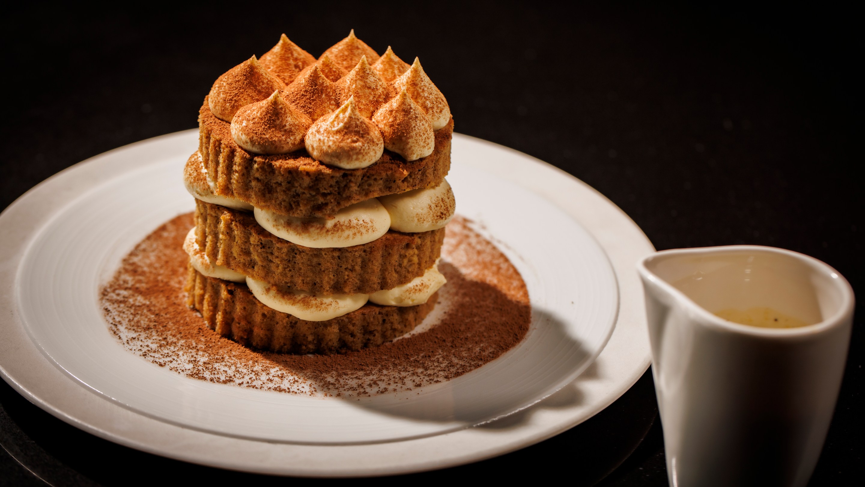 Tiramisu dessert layered with mascarpone and cocoa on a white round plate and a dish of sauce next to it at The Saint Dallas