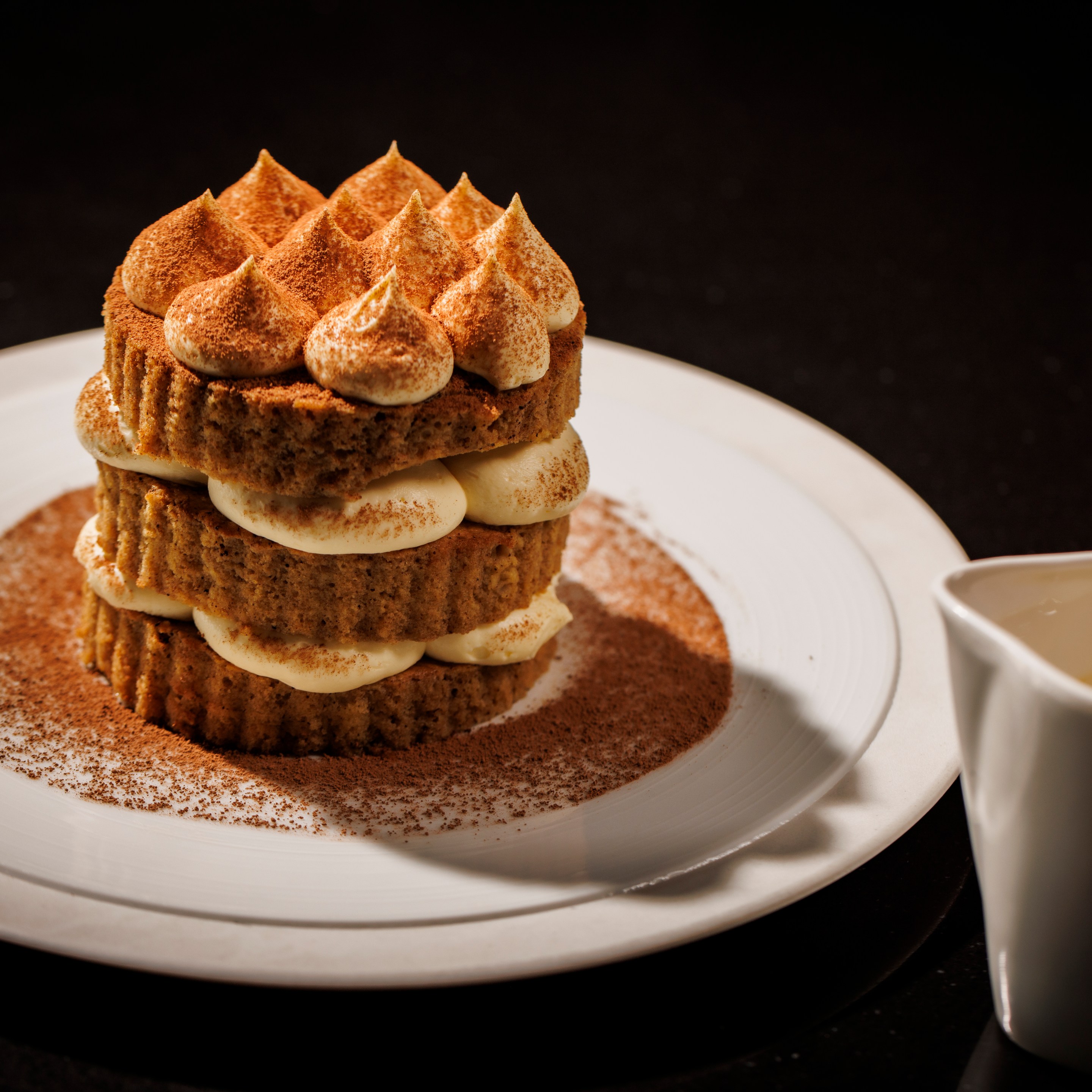 Tiramisu dessert layered with mascarpone and cocoa on a white round plate and a dish of sauce next to it at The Saint Dallas