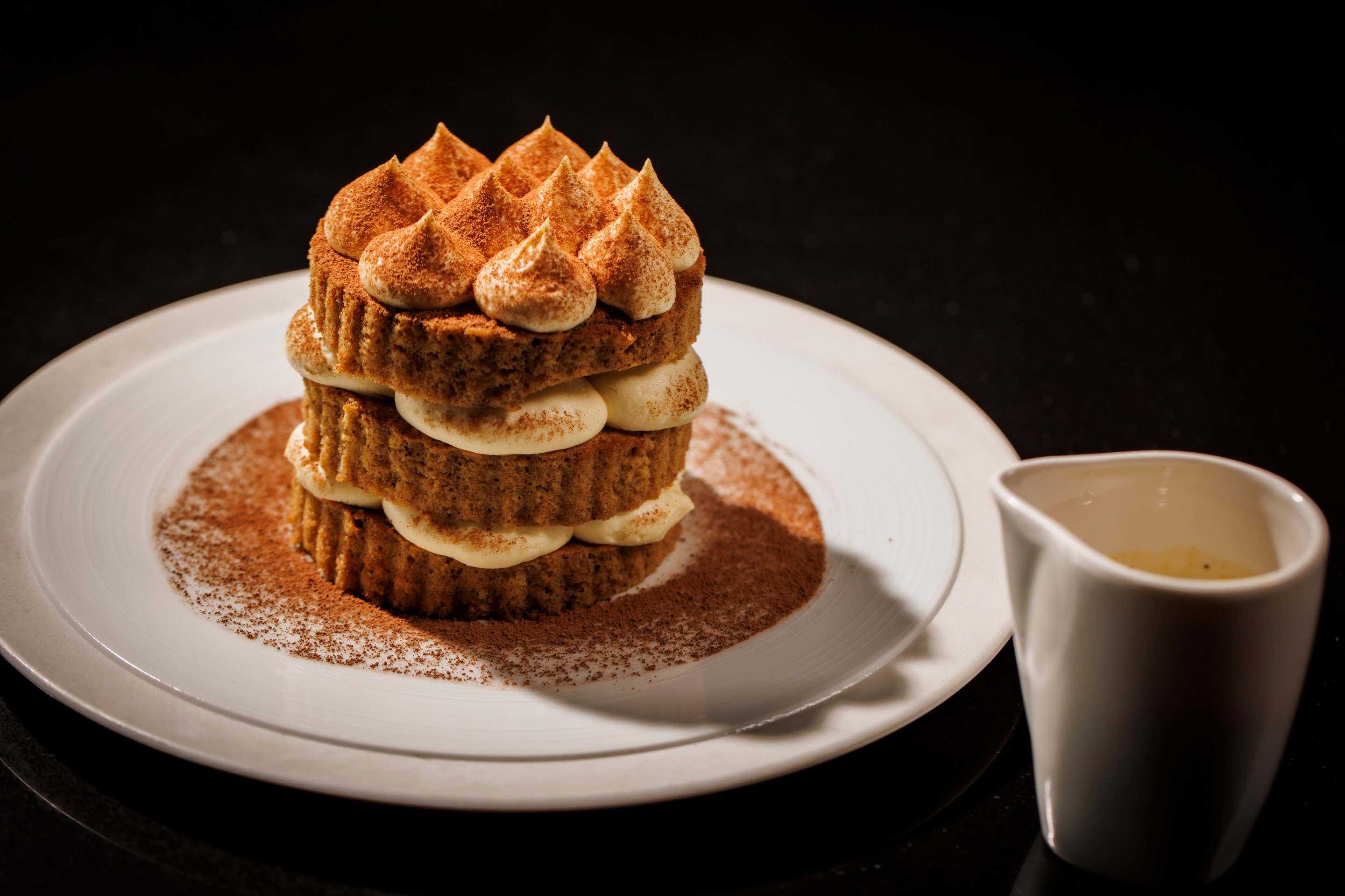 Tiramisu dessert layered with mascarpone and cocoa on a white round plate and a dish of sauce next to it at The Saint Dallas