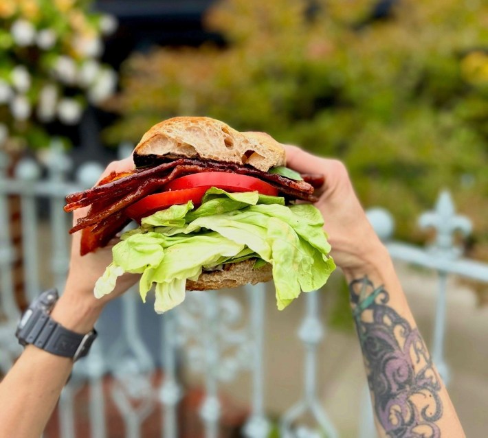 Person holding sandwich made with focaccia bread, bacon, tomato, and lettuce