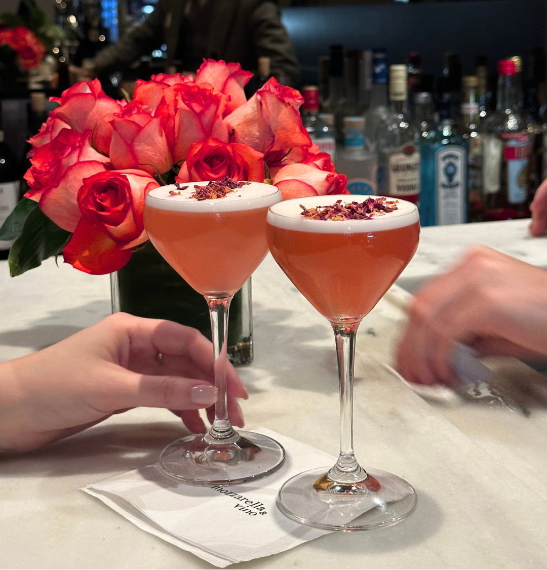 Two pink Rose Kiss cocktails garnished with rose petals on a marble bar at Mozzarella & Vino with red roses in the background.