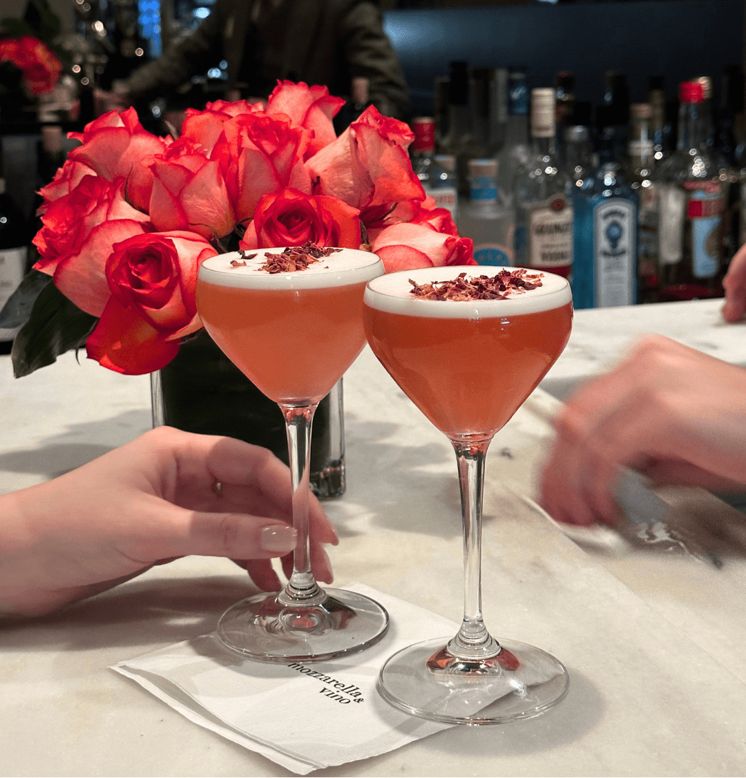 Two pink Rose Kiss cocktails garnished with rose petals on a marble bar at Mozzarella & Vino with red roses in the background.