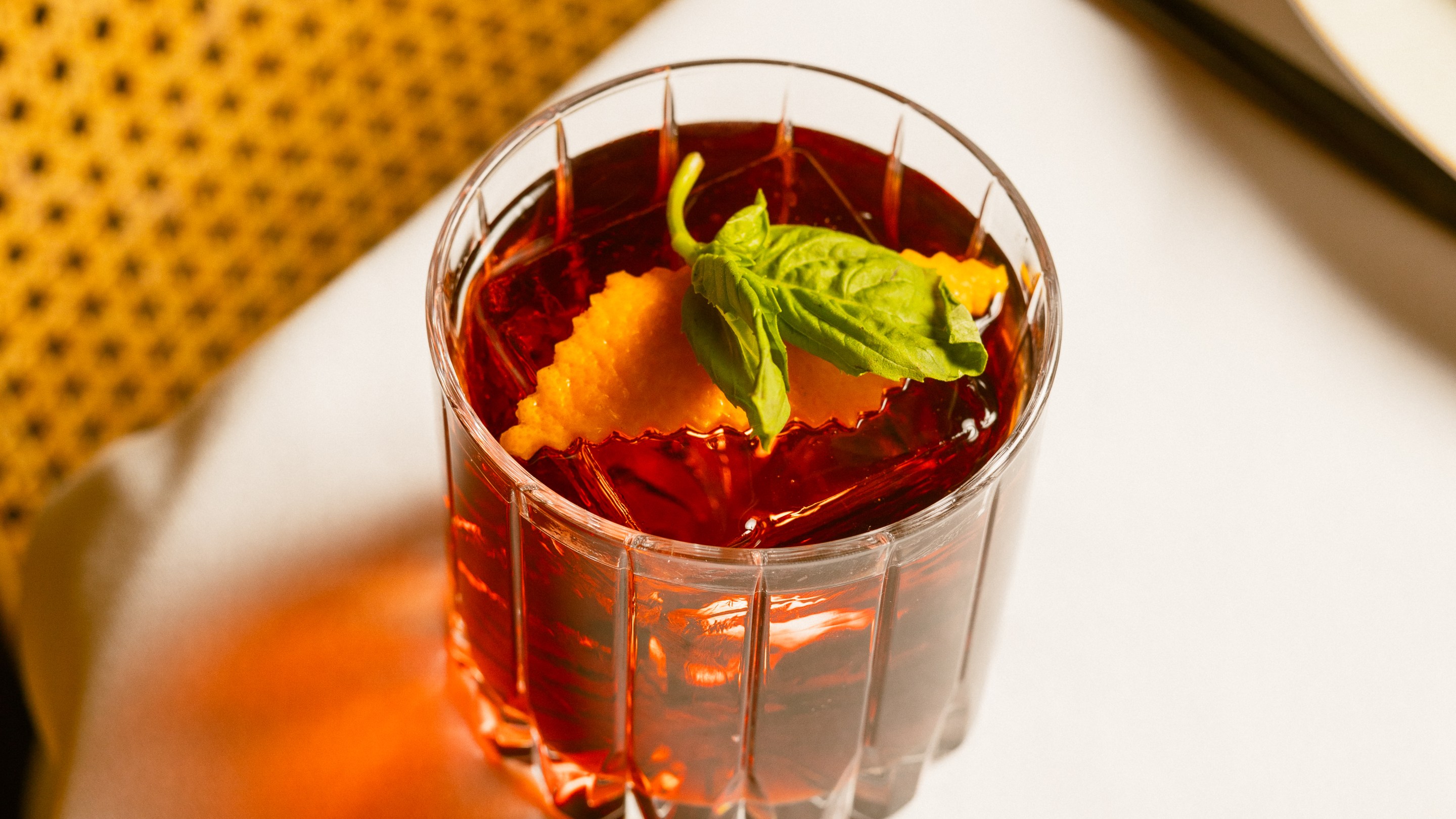 Close-up of a negroni cocktail in a cut-glass tumbler with orange garnish and basil leaf.