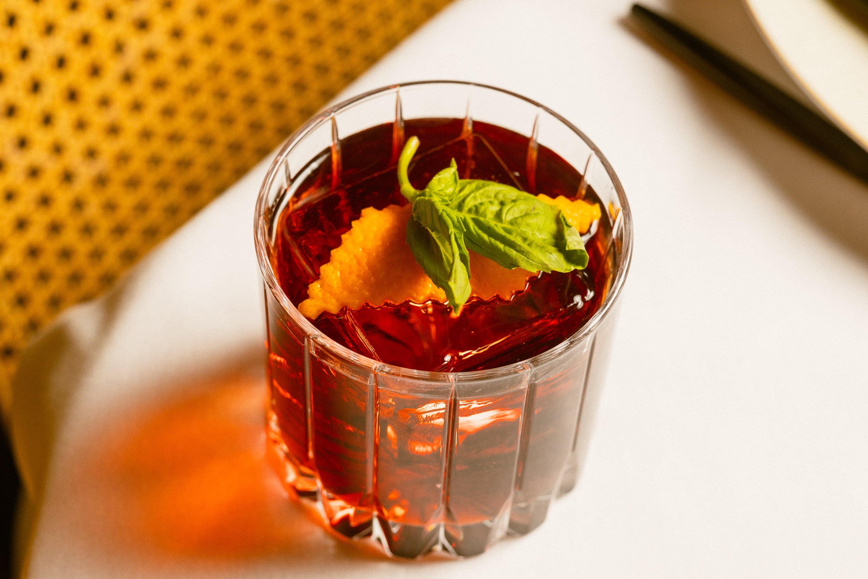 Close-up of a negroni cocktail in a cut-glass tumbler with orange garnish and basil leaf.