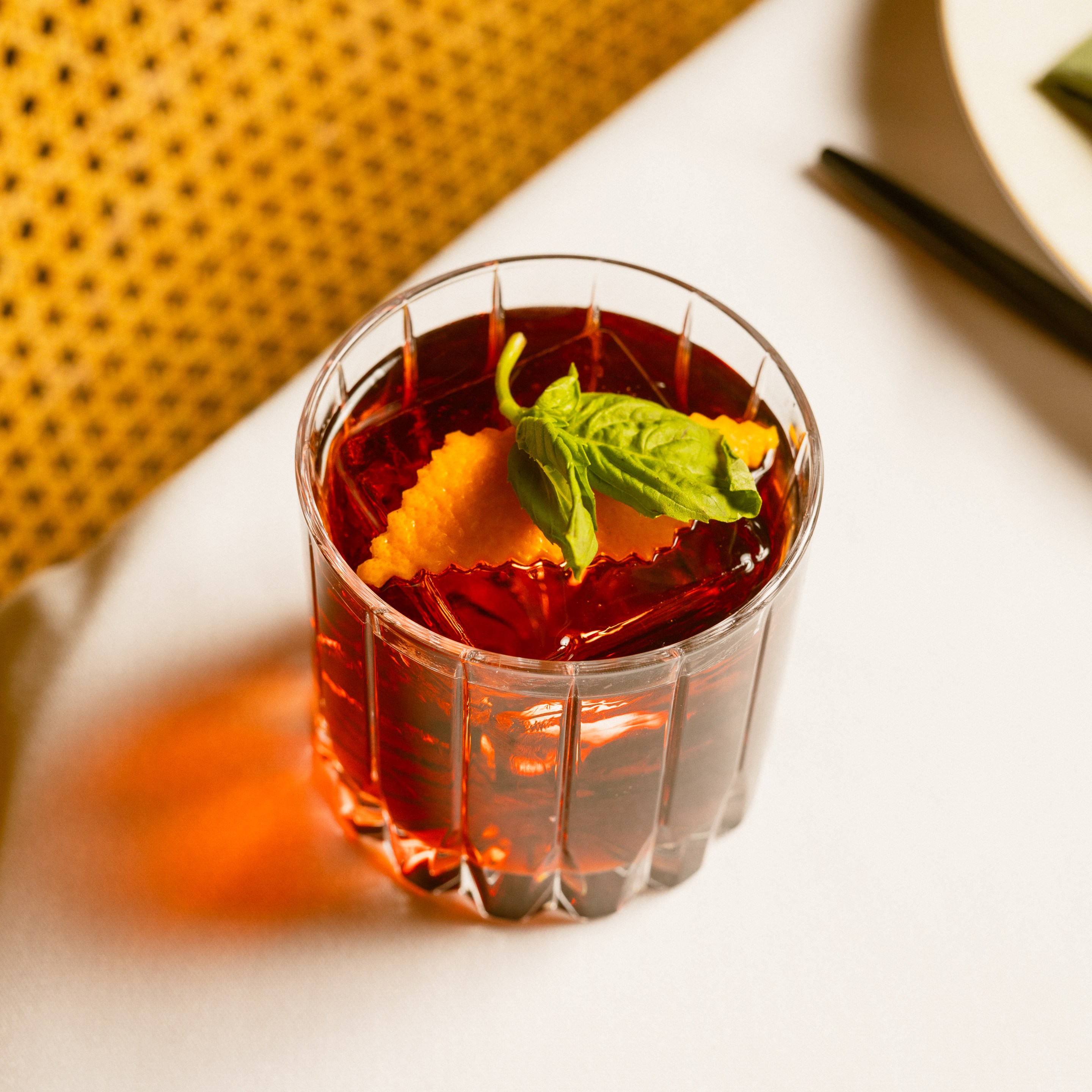 Close-up of a negroni cocktail in a cut-glass tumbler with orange garnish and basil leaf.