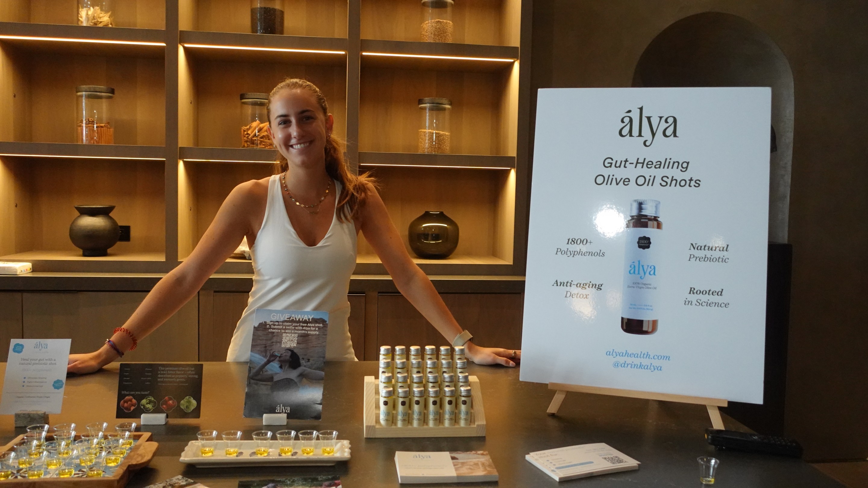Founder Maya Ayed standing behind a display of Alya Health olive oil shots at an event