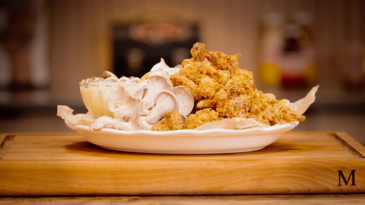 Crispy popcorn mushrooms served with dipping sauce, a simple preparation that emphasizes texture and flavor, served in a white dish on a wooden cutting board.