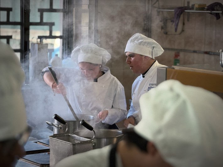 Culinary students cooking at a stove with steam rising from pots in a professional kitchen.