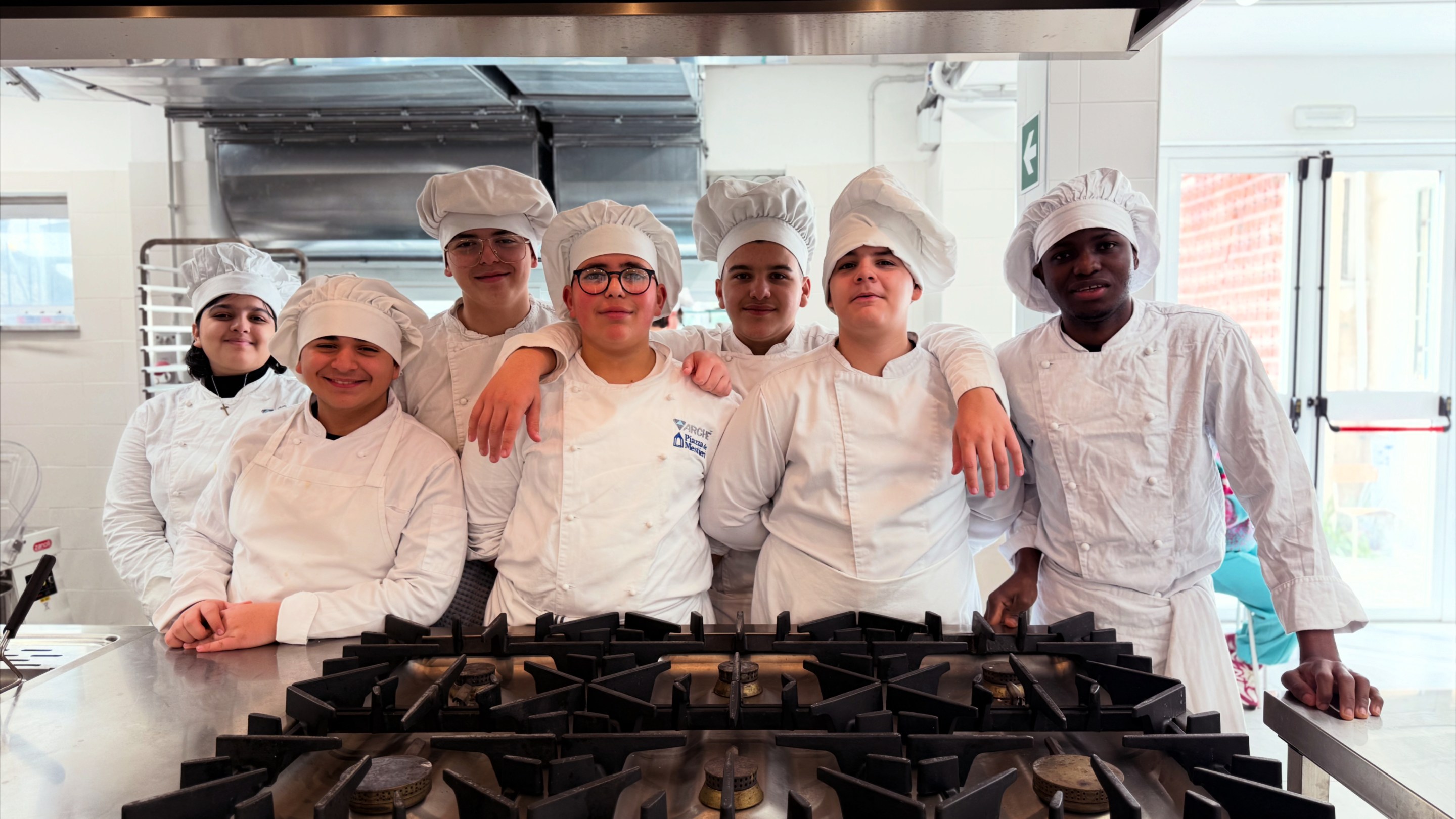 Group of young culinary students in chef uniforms standing together in a professional kitchen.