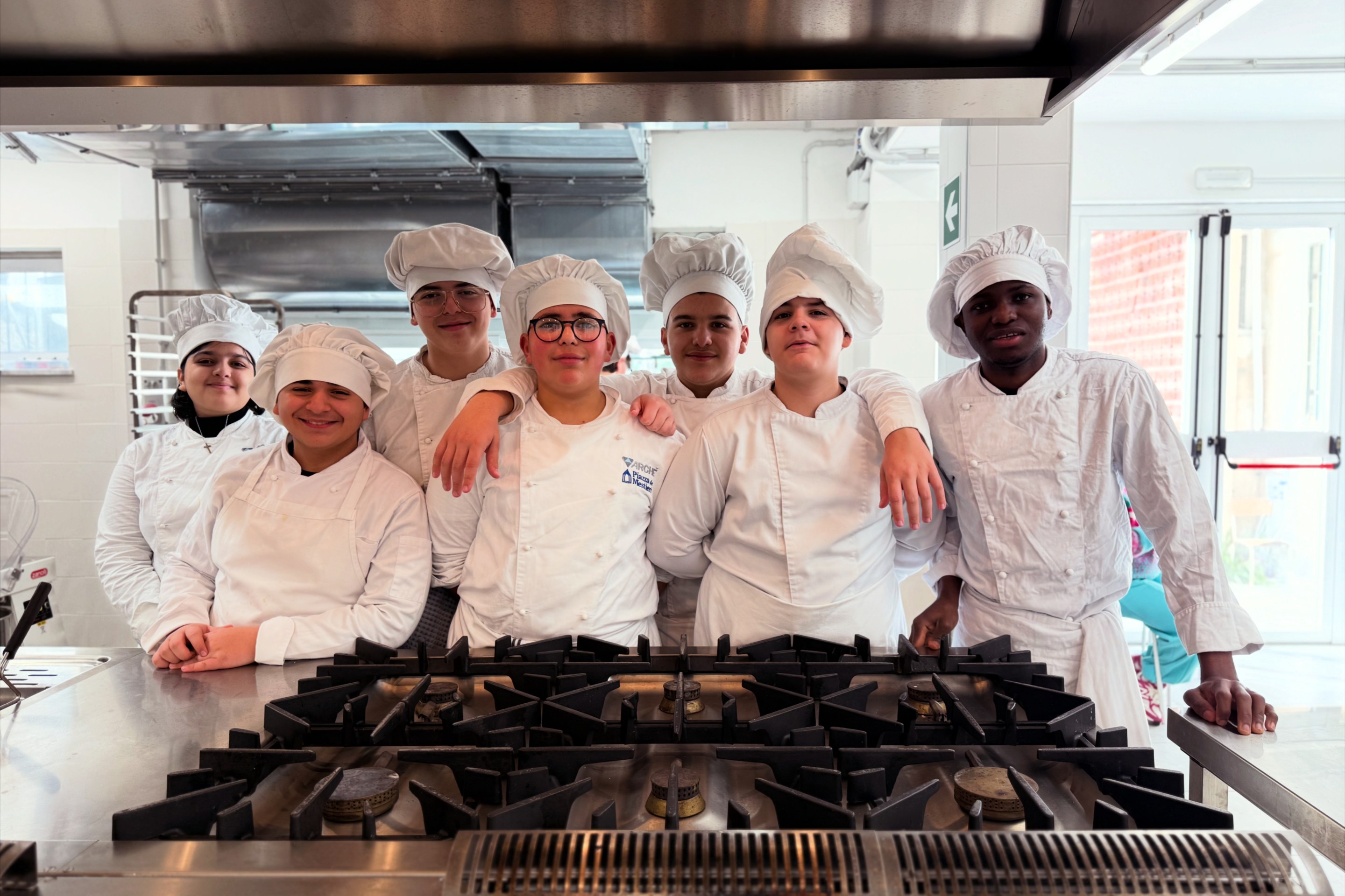 Group of young culinary students in chef uniforms standing together in a professional kitchen.
