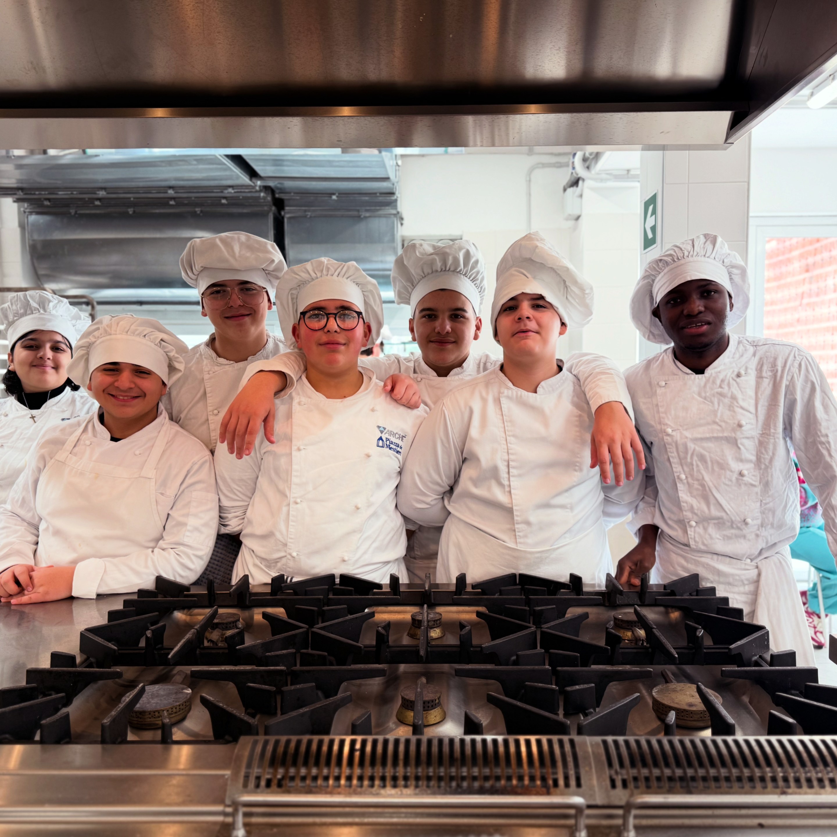 Group of young culinary students in chef uniforms standing together in a professional kitchen.