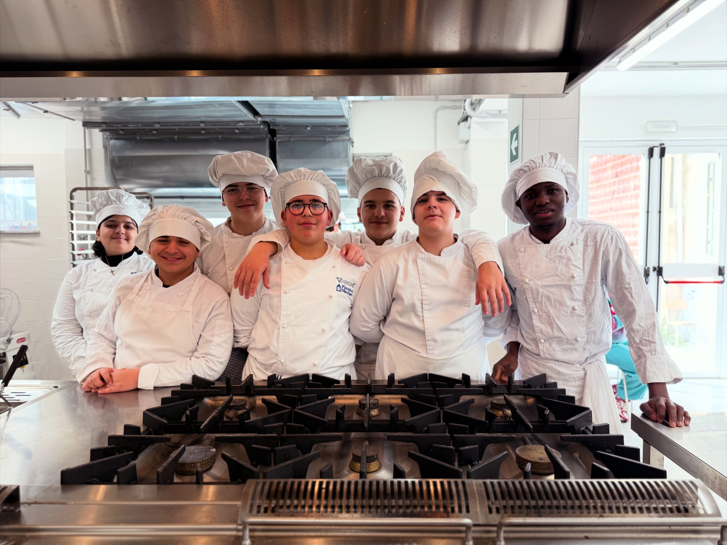 Group of young culinary students in chef uniforms standing together in a professional kitchen.