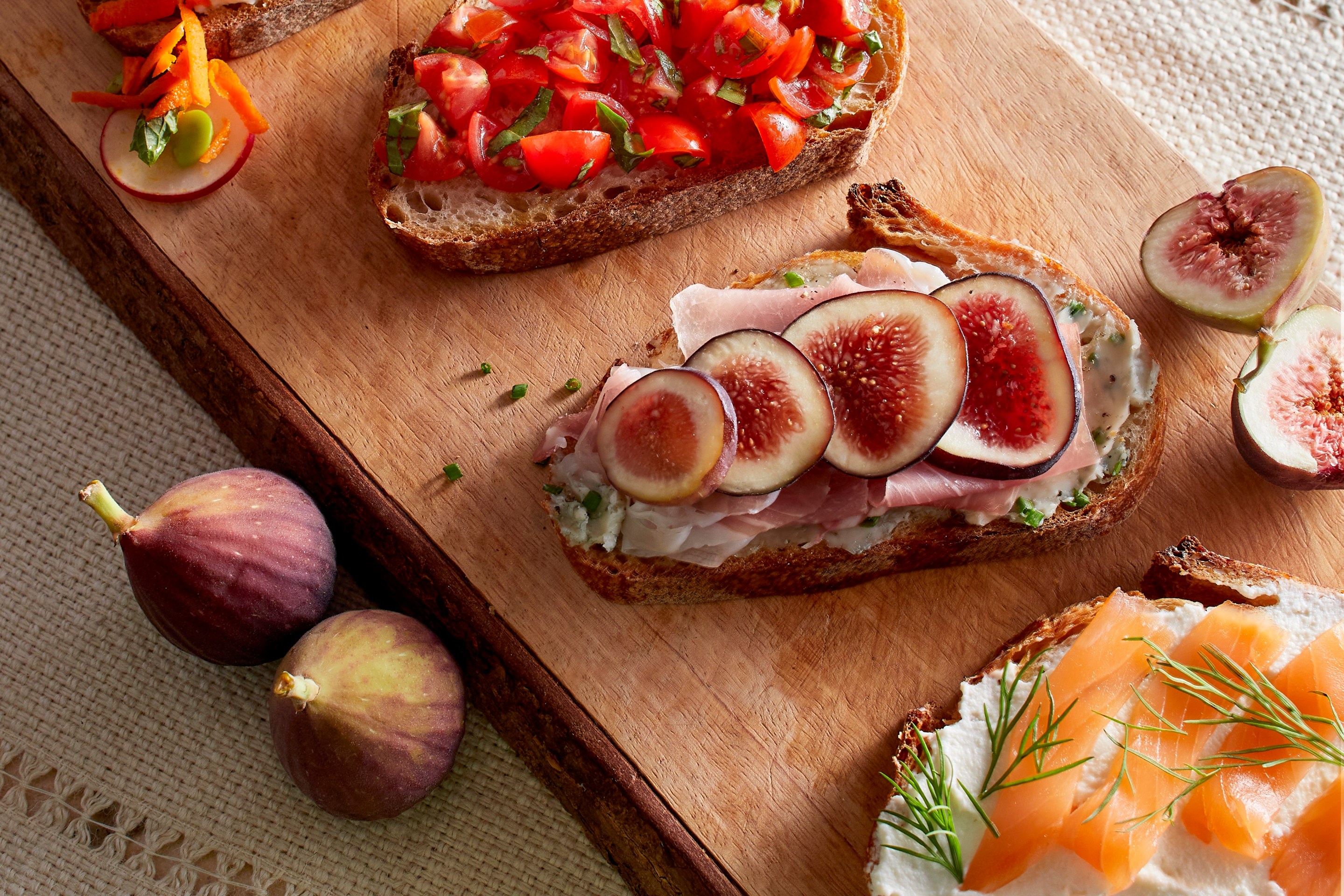 A rustic bruschetta board topped with fresh tomatoes, basil, burrata, prosciutto, olives, roasted vegetables, and toasted bread