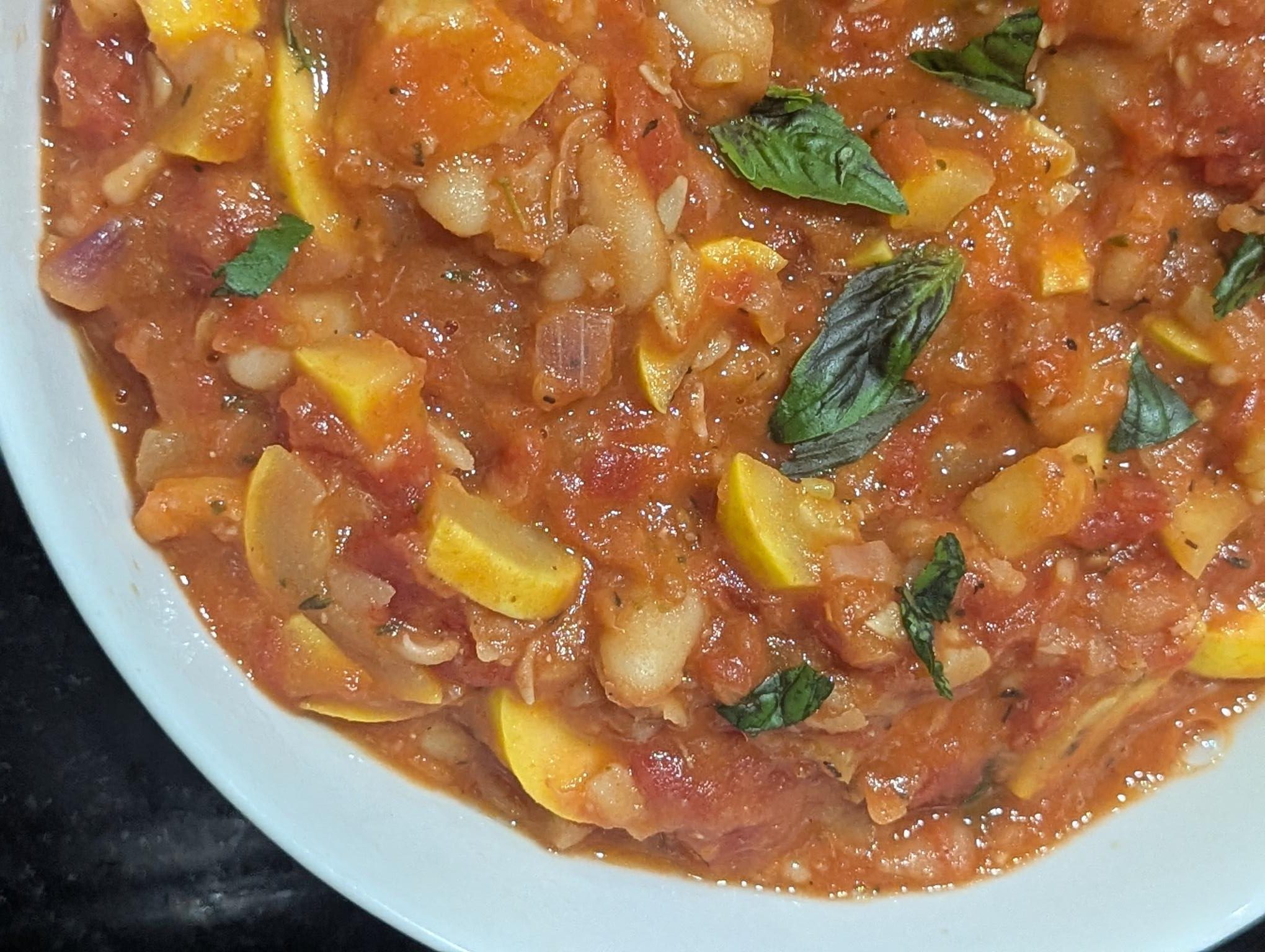Close-up of homemade yellow squash stew with tomatoes and basil in a white bowl.