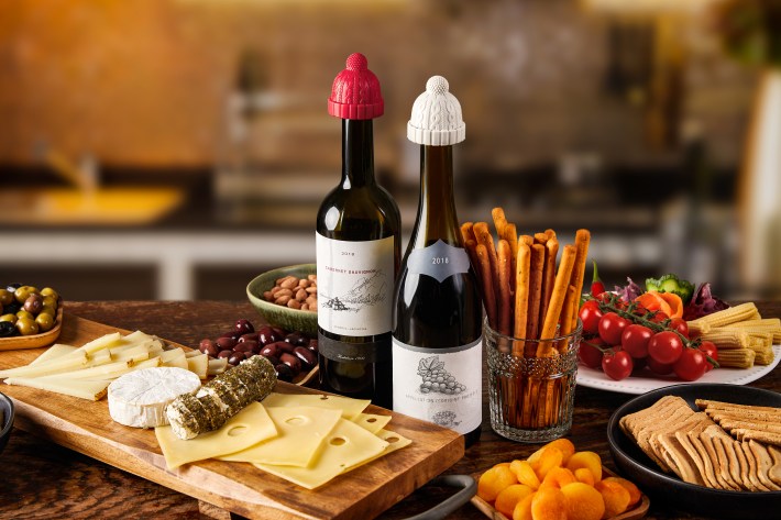 Wine bottles fitted with red and white winter hat bottle stoppers beside a cheese and appetizer spread