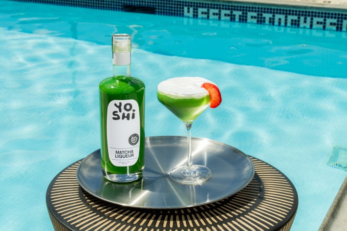 Bottle of YOSHI Matcha Liqueur with a green Matcha Martini beside a swimming pool