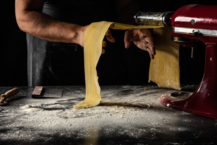 Hands guide a sheet of fresh pasta dough through a roller over a floured counter.