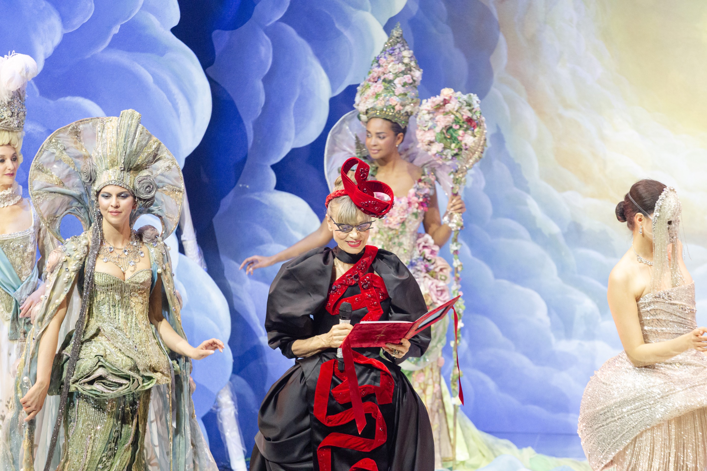Antonia Sautter holds a microphone onstage, surrounded by performers in ornate costumes.