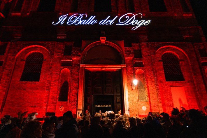 Guests in Carnival attire gather outside a red-lit entrance marked Il Ballo del Doge.