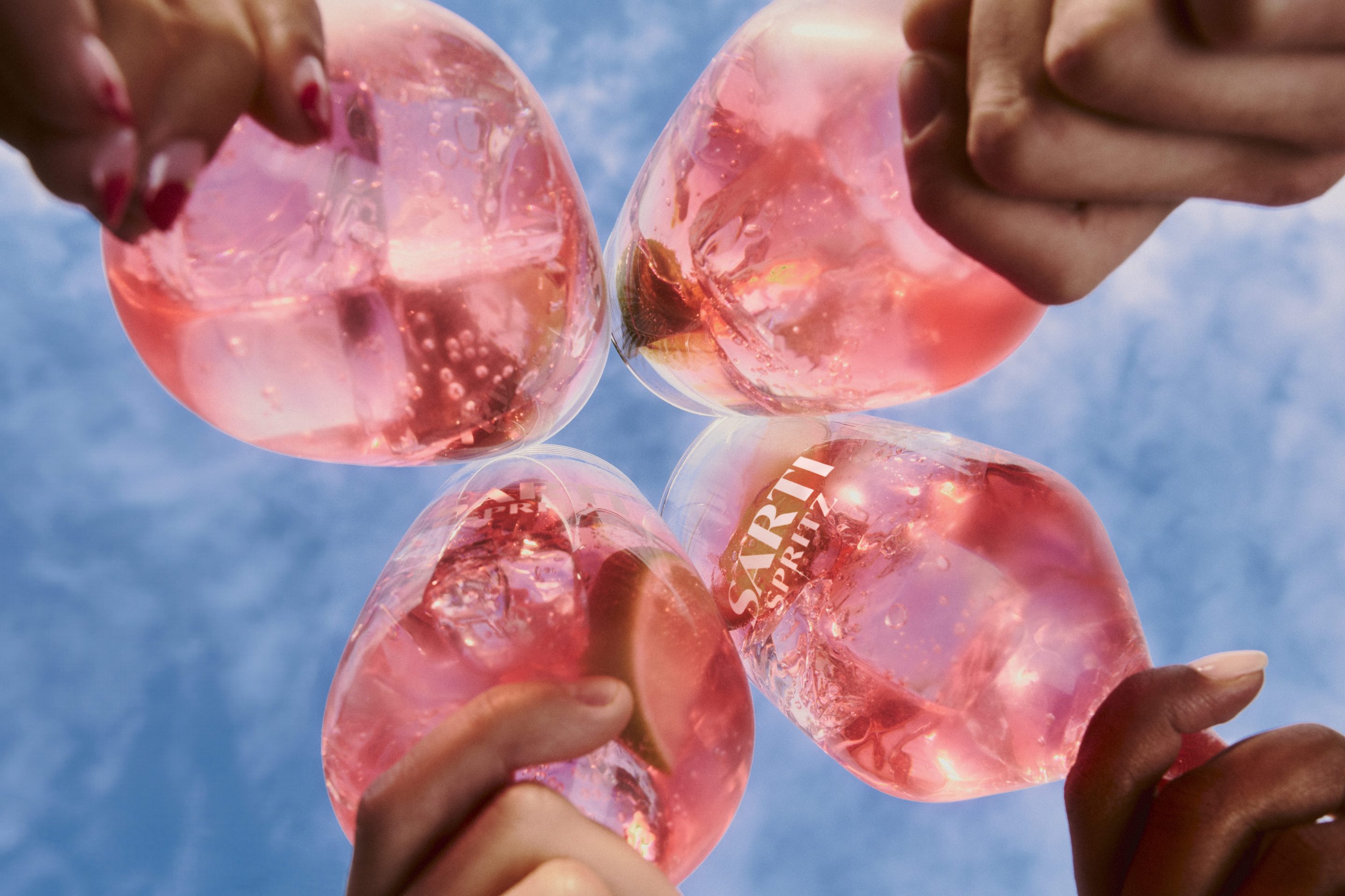 Four hands toast with pink Sarti Spritz cocktails against a blue sky.